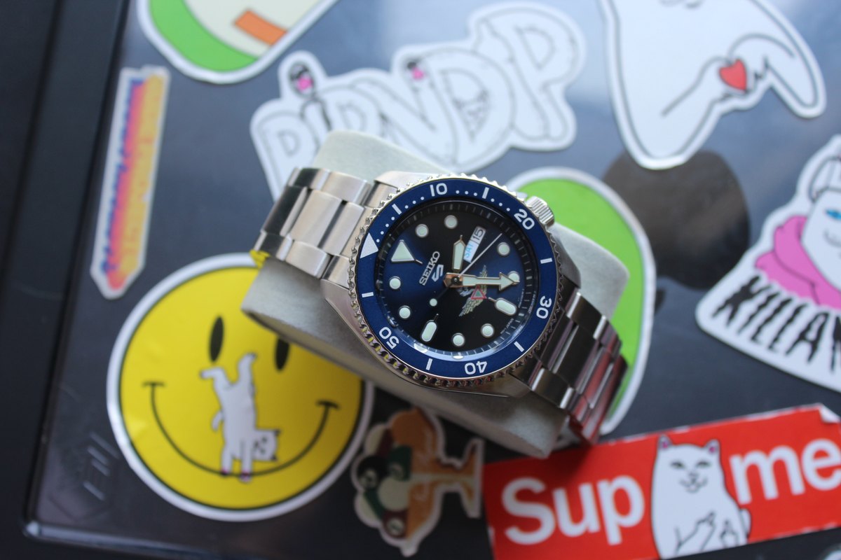 El reloj apoyado sobre la tapa de una portátil cubierta de calcomanías.