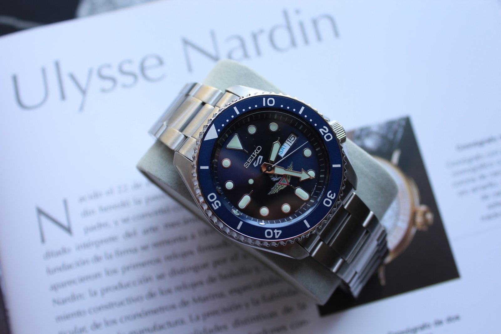 Reloj de acero con esfera y bisel azules, con un emblema alado impreso en la carátula.