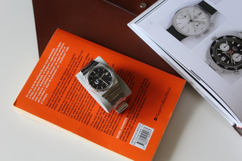 El reloj apoyado sobre un libro naranja, junto a una revista abierta.